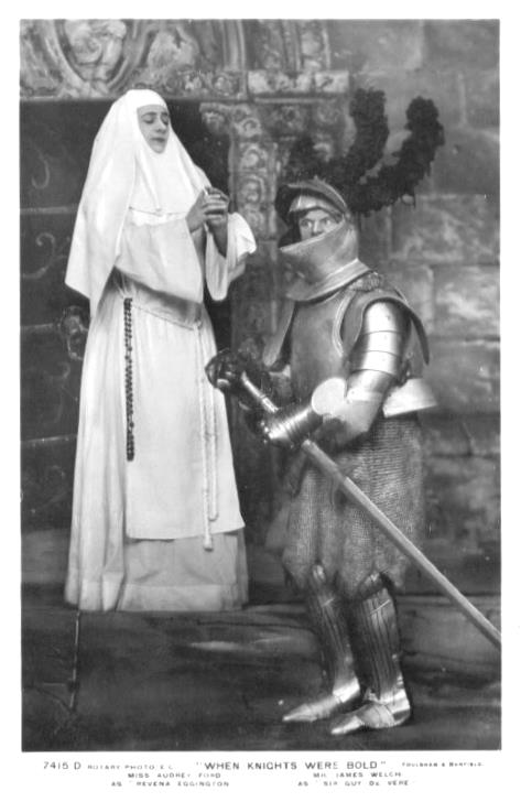 A puzzled James Welch (as Sir Guy, dressed in a full suit of armor) stands in front of a praying Audrey Ford (as Revena Eggington, in a white nun’s habit).